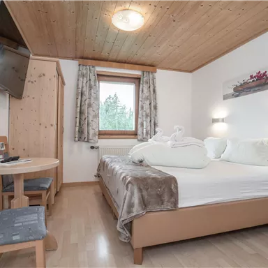 A cozy hotel room with a double bed, a table, and chairs. The room features a wooden ceiling and a large television.