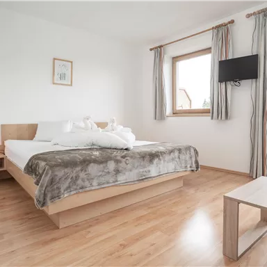 A bright, modern bedroom with a large bed and wooden floor. There are large windows with curtains and a TV on the wall.