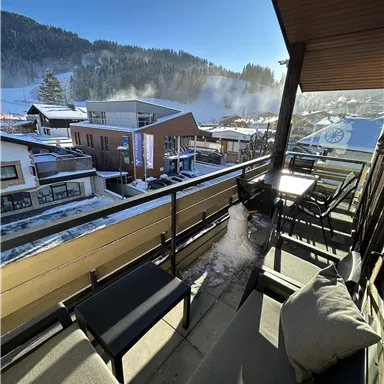 Ein Balkon mit Sitzgelegenheiten und Blick auf ein verschneites Dorf. Im Hintergrund sind Berge und blauer Himmel zu sehen.
