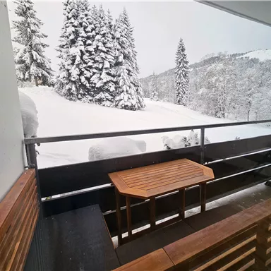 A balcony with a wooden deck table and seating options, surrounded by snow-covered trees. The landscape appears wintry and peaceful.