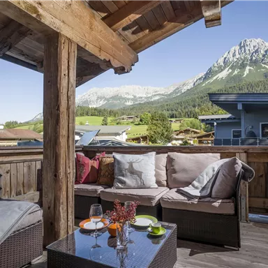 A cozy balcony with seating and a view of majestic mountains. The clear sky and the rays of sunshine create an inviting atmosphere.