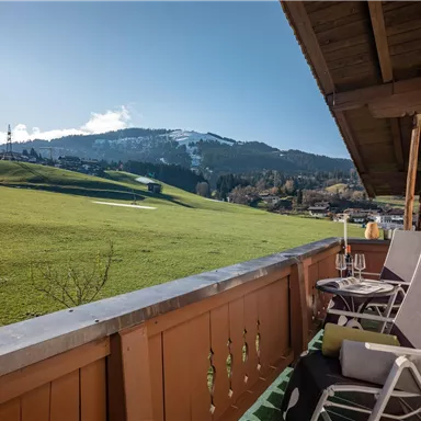 Ein Balkon mit zwei Liegestühlen und Blick auf grüne Wiesen und Berge. Die Sonne scheint und der Himmel ist blau mit einigen Wolken.