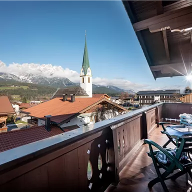 Ein Balkon mit Blick auf eine malerische Berglandschaft. Im Hintergrund sieht man eine Kirche mit einem spitzen Turm und das typische alpine Dorfleben.