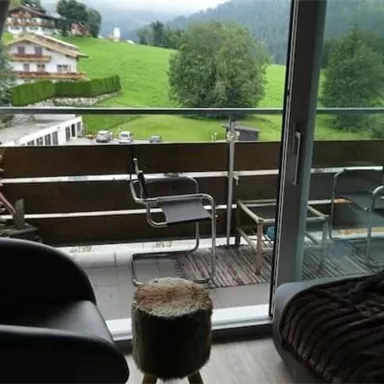 A balcony with two chairs and a small stool. In the background, green meadows and mountains can be seen.