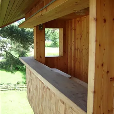 Eine Holzveranda mit einem offenen Fenster und einer breiten Ablagefläche. Der Blick fällt auf eine grüne Wiese und Bäume im Hintergrund.