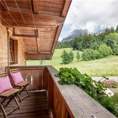 Ein gemütlicher Balkon aus Holz mit zwei Stühlen und Kissen. Im Hintergrund erstreckt sich eine grüne Wiese und bewaldete Berge.