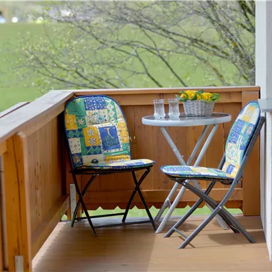A small balcony with two colorful chairs and a table. On the table, there are a few glasses of water and a flower bowl.