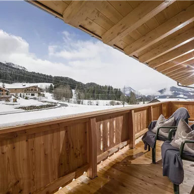 Ein gemütlicher Balkon mit zwei Stühlen und einem wunderschönen Blick auf eine verschneite Landschaft. Im Hintergrund sind Berge und Hütten zu sehen.