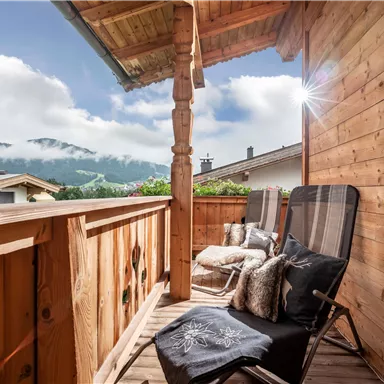 A cozy wooden balcony with two chairs and a view of the mountains. The clouds are moving across the landscape while the sun is shining.