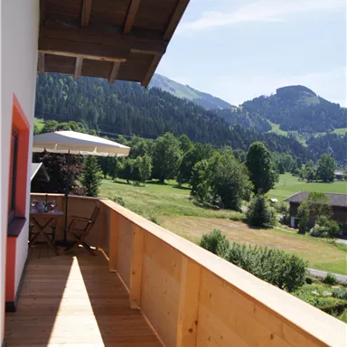 A beautiful balcony with a wooden railing and a view of green meadows and mountains. The surrounding landscape is sunny and inviting.