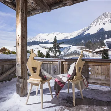 A balcony with two wooden chairs and a blanket. In the background, snow-covered mountains and a clear sky can be seen.