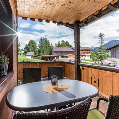 A cozy balcony with a table and chairs. In the background, green meadows and mountains can be seen under a clear sky.