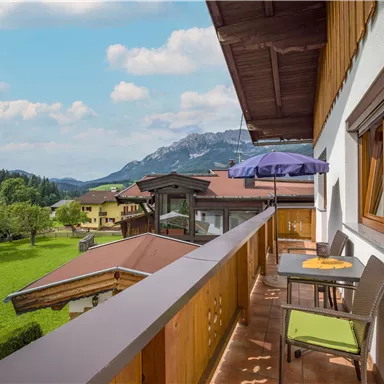 A beautiful balcony with a view of the mountains and the green landscape. There is a table and chairs, and a sun umbrella provides shade.