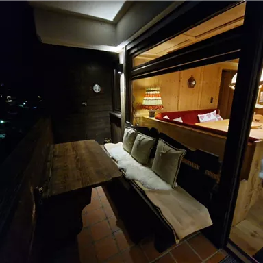 Ein gemütlicher Balkon mit einer Holzbank und Kissen. Im Hintergrund sieht man einen warm beleuchteten Wohnbereich.