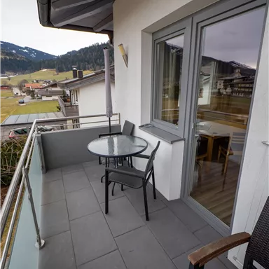 Ein kleiner Balkon mit einem runden Tisch und vier Stühlen. Im Hintergrund sind Berge und eine ruhige Landschaft zu sehen.