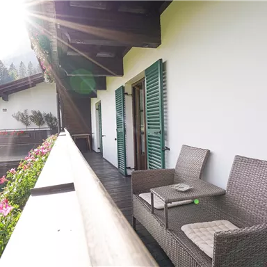 Ein sonniger Balkon mit modernem Rattanmöbel und bunten Blumen. Die Fensterläden sind grün und die Umgebung ist ländlich und schön.