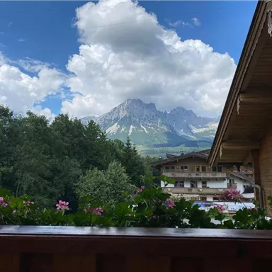 Eine malerische Berglandschaft mit majestätischen Bergen und dichten Wäldern. Im Vordergrund blühen farbige Blumen auf einem Balkon.