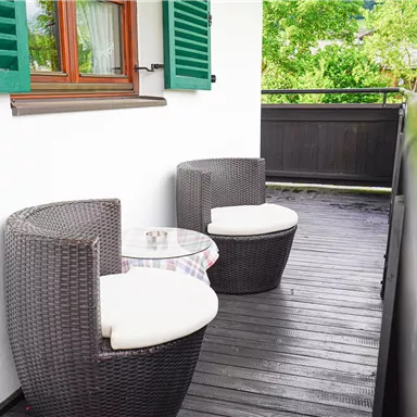Ein gemütlicher Balkon mit zwei Rattan-Stühlen und einem kleinen Tisch. Im Hintergrund sind grüne Pflanzen und Fenster mit Holzläden.