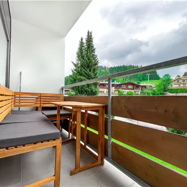 Ein moderner Balkon mit Holzmöbeln und bequemen Sitzkissen. Im Hintergrund sind grüne Hügel und Wolken am Himmel sichtbar.