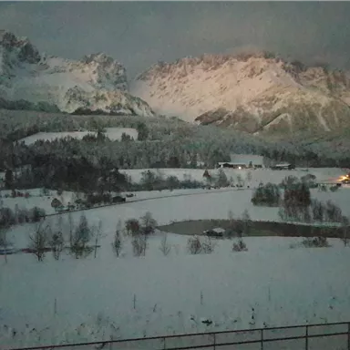 A snowy landscape with high mountains in the background. The surroundings are quiet and some lights can be seen in the distance.