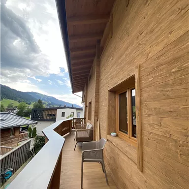 A spacious balcony with wooden paneling and a view of the mountains. In the background, more houses and a cloudy sky can be seen.