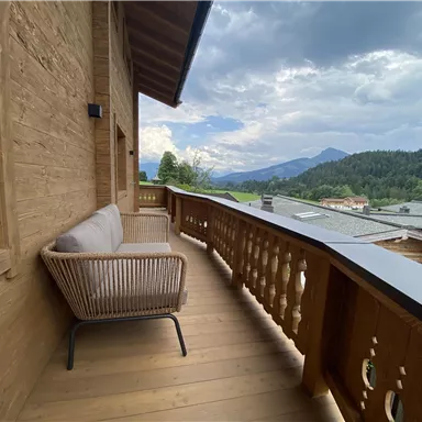 A wooden balcony with a cozy chair and a view of green hills and mountains. The sky is slightly cloudy, providing a relaxed atmosphere.