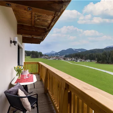 A beautiful balcony with a wooden railing and a view of a green landscape. In the foreground, there is a table with a tablecloth and flowers, as well as a chair.
