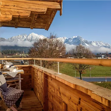 Ein Balkon mit Holzverkleidung und zwei Stühlen. Im Hintergrund sind schneebedeckte Berge und ein klarer Himmel zu sehen.