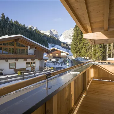 A beautiful balcony with a wooden railing and a view of a snowy valley. In the background, houses and snow-covered mountains are visible.