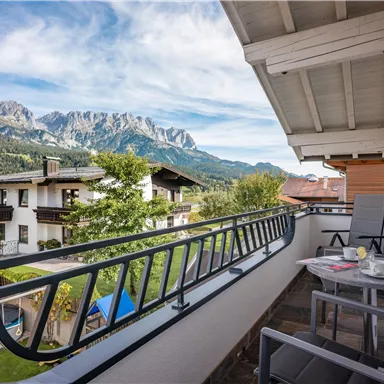 Eine gemütliche Balkonansicht mit Stühlen und einem Tisch. Im Hintergrund sind beeindruckende Berge und grüne Bäume zu sehen.