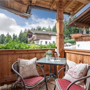 A cozy balcony with two chairs and a small table. In the background, green trees and traditional houses can be seen.