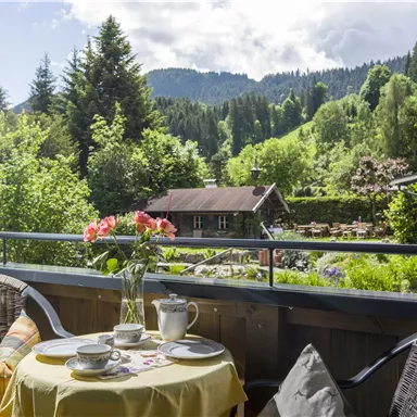 Ein gemütlicher Balkon mit gedecktem Tisch und Blick auf grüne Berge. Auf dem Tisch stehen eine Blumenvase, Tassen und Geschirr.
