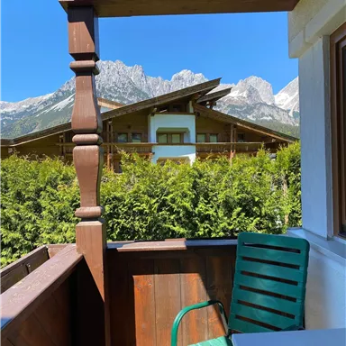Blick von einem Balkon auf eine grüne Hecke und beeindruckende Berge im Hintergrund. Die Sonne scheint bei strahlend blauem Himmel.