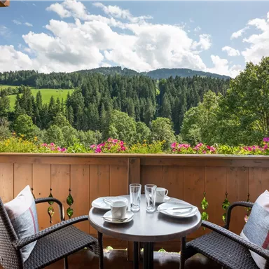 Ein Balkon mit einem kleinen Tisch und zwei Stühlen. Im Hintergrund sind grüne Hügel und Wälder unter einem hellen Himmel zu sehen.