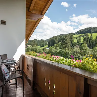 Ein hübscher Balkon mit zwei Stühlen und einem Tisch, umgeben von bunten Blumen. Im Hintergrund erstreckt sich eine grüne Landschaft unter einem klaren Himmel.