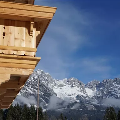 A traditional wooden balcony overlooking snow-capped mountains. The sky is blue and clear, surrounded by trees.