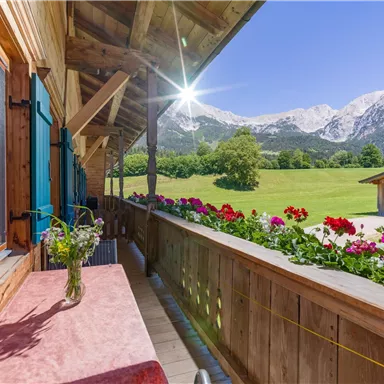 Eine sonnige Terrasse mit bunten Blumen und Blick auf die Berge. Der Himmel ist klar und die Landschaft ist grün.