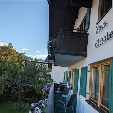 Ein charmantes Haus mit dem Namen "Haus Elisabeth" und grünen Fensterläden. Im Vordergrund sieht man einen Balkon mit Blick auf einen Garten und die umliegende Landschaft.
