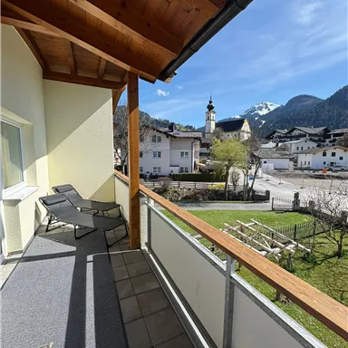 Ein Balkon mit Liegen und Blick auf einen malerischen Ort in den Bergen. Die Umgebung ist grün und der Himmel ist klar.