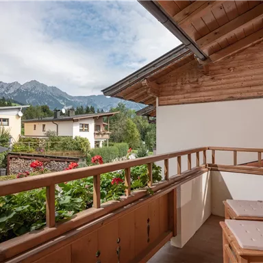 Ein gemütlicher Balkon mit Holzgeländer und zwei Stühlen. Im Hintergrund sind Berge und eine grüne Landschaft zu sehen.