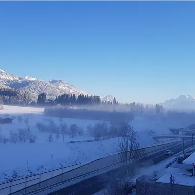 A winter landscape with snow-covered mountains and a clear blue sky. In the foreground, there is a road and some buildings visible.
