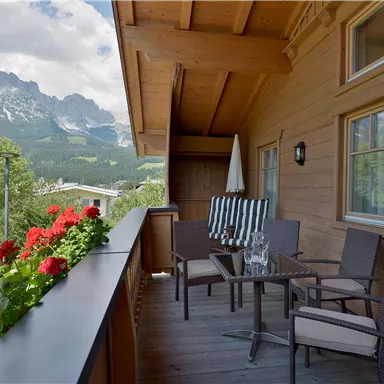 Ein gemütlicher Balkon mit Stühlen und Tisch, umgeben von blühenden Geranien. Im Hintergrund sind majestätische Berge und ein blauer Himmel sichtbar.