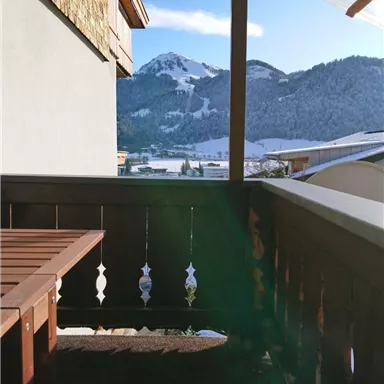 Ein gemütlicher Balkon mit Holzgeländer und Tisch. Im Hintergrund sind schneebedeckte Berge und ein blauer Himmel zu sehen.