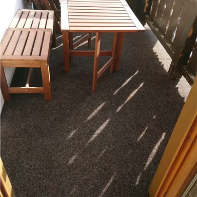 Ein kleiner Balkon mit einem Tisch und zwei Stühlen. Der Boden ist mit einem Teppich bedeckt, und es ist Sonnenschein zu sehen.