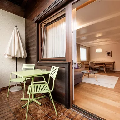 A cozy balcony with a small table and chairs in green. In the background, a bright living space can be seen.