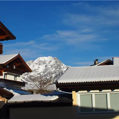 Eine winterliche Landschaft mit schneebedeckten Dächern und Bergen im Hintergrund. Der Himmel ist klar und blau.
