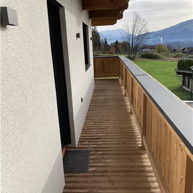 Ein Holz-Balkon mit Blick auf eine grüne Wiese und Berge im Hintergrund. Die Wand ist hell und der Himmel ist teils bewölkt.