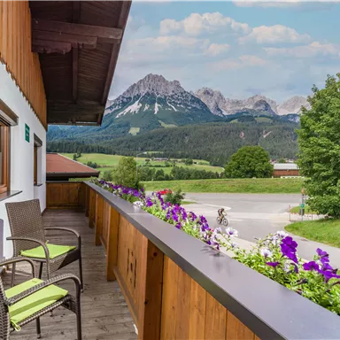 A cozy balcony with two chairs and beautiful flowers, offering a view of the majestic mountains and the green landscape. The sky is clear and nature is idyllic.