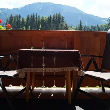 Ein gemütlicher Balkon mit zwei Stühlen und einem kleinen Tisch. Im Hintergrund ist eine schöne Berglandschaft und Bäume zu sehen.