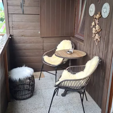 A cozy balcony with two chairs and a small table. The walls are made of wood, and a soft blanket is lying on one of the chairs.